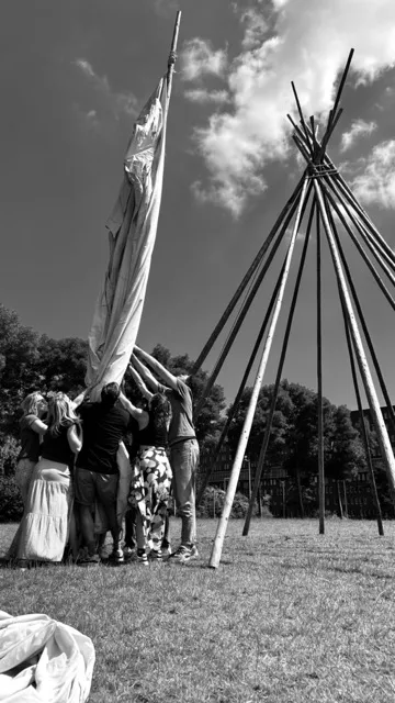 een groep mensen die het doen van een immens grote tip tent proberen te plaatsen op het houten geraamte. Het gaat hier om Teamwork.