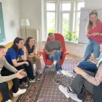 een foto van een groep mensen die een xworkshop volgen. # mensen zitten op een gele bank, 1 zit op een rode stoel en 2 zitten op een houten stoel. De Change Agent die de workshop geeft staat voor de groep en naast haar staat en flip-over.