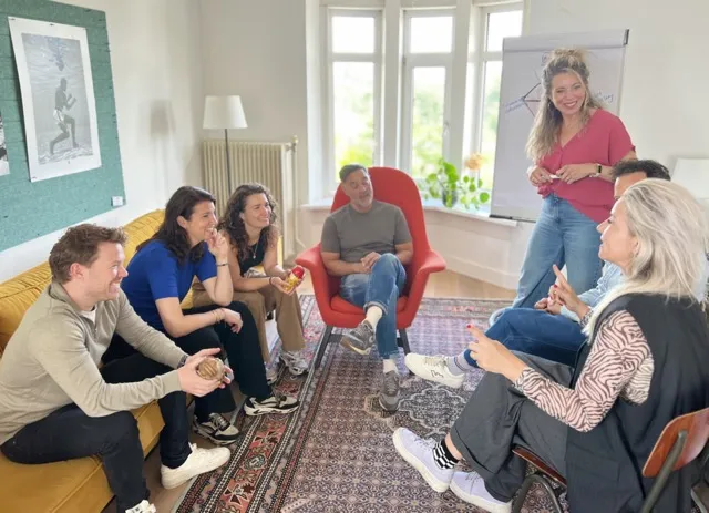 een foto van een groep mensen die een xworkshop volgen. # mensen zitten op een gele bank, 1 zit op een rode stoel en 2 zitten op een houten stoel. De Change Agent die de workshop geeft staat voor de groep en naast haar staat en flip-over.